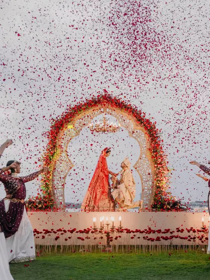 A truly magical moment as Maheswari and Basu exchange garlands under a shower of red rose petals, framed by dancers and a stunning floral arch.