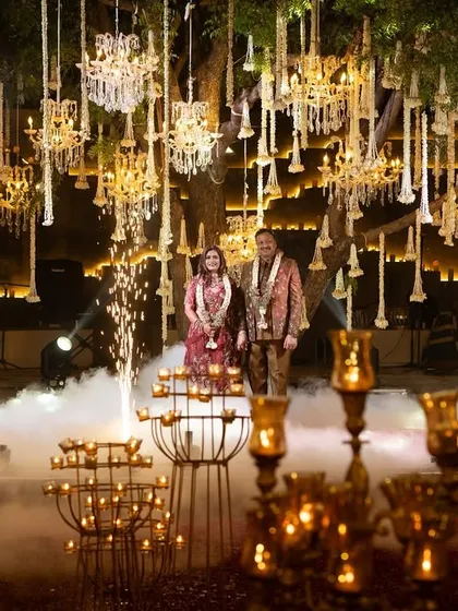The couple stands on stage during their 25th-anniversary celebration, framed by cold pyrotechnics. The backdrop of hanging chandeliers and florals creates a truly spectacular and grand setting.