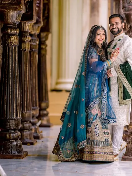A happy couple poses in a corridor of wooden pillars. The bride's beautiful blue lehenga and the groom's elegant sherwani create a timeless, royal look.