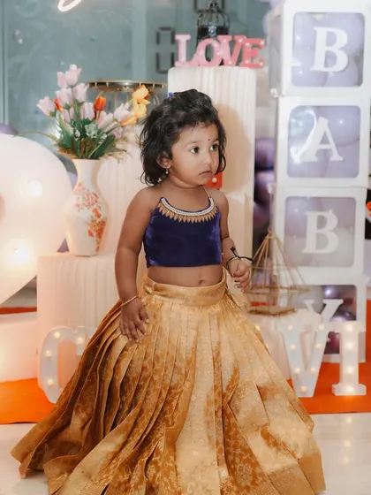 This little client is wearing a custom designer kids' lehenga choli. The outfit pairs a rich navy blue velvet top with a beautifully pleated golden silk brocade skirt, creating a stunning traditional look for her birthday celebration.