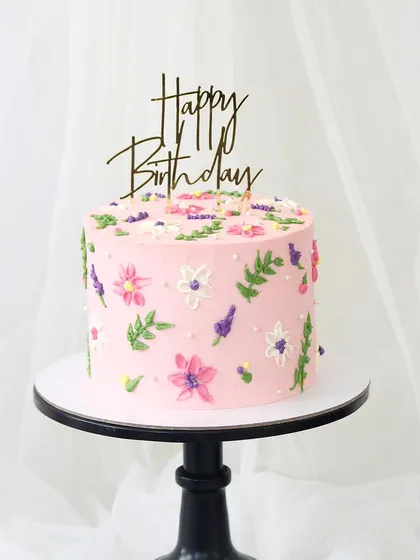 A lovely pink birthday cake with a hand-piped wildflower meadow design. The colorful flowers and green leaves create a fresh and charming look, perfect for a spring or summer celebration.