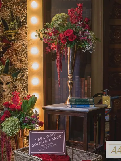 A cozy corner from our speakeasy-themed event, featuring a tall floral arrangement in reds and greens, vintage books, and a playful sign inviting guests to "Save your soles & dance!". These small details help build the narrative of the theme.
