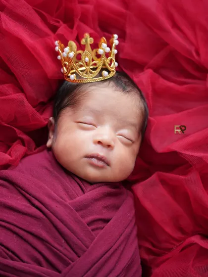 A detailed close-up of a newborn wearing a delicate pearl and gold crown, swaddled in red. This shot emphasizes the royal theme and the baby's peaceful slumber.