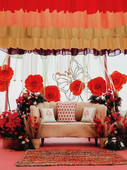A closer look at the vibrant Mehendi stage, featuring bold red paper flowers, a wireframe butterfly motif, and layered, colorful ceiling drapes.