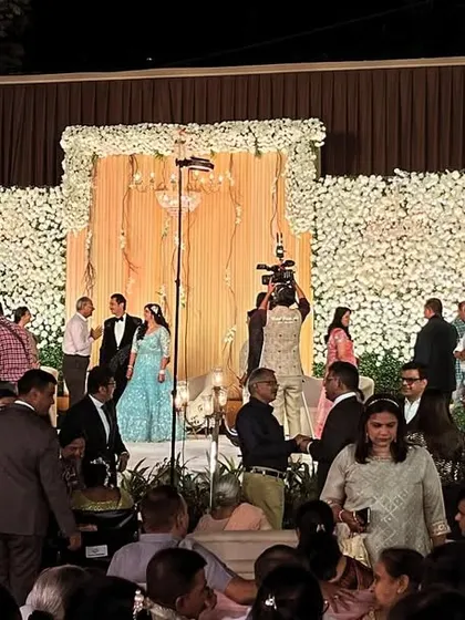 A candid shot of a busy wedding reception at The Acres Club. The stage is decorated with a wall of white flowers, creating a beautiful focal point for the event.