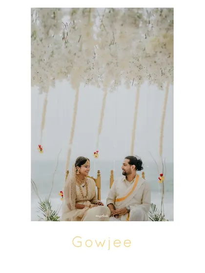A beautiful portrait of the couple seated at their beach wedding mandap, under a canopy of hanging white flowers.