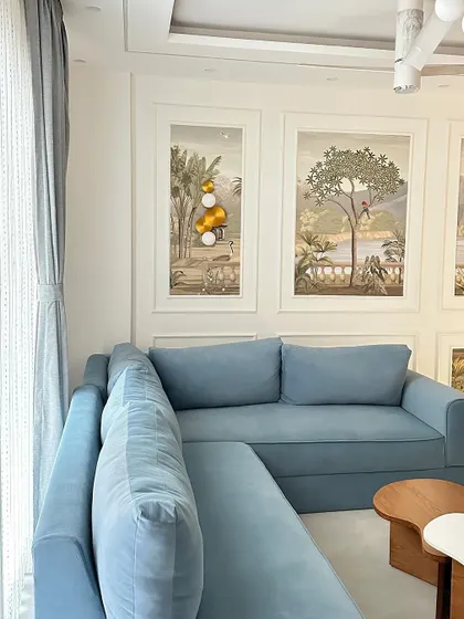 A cozy living room featuring a custom light blue L-shaped sofa. The walls are adorned with scenic wallpaper panels, creating a serene and artistic backdrop.