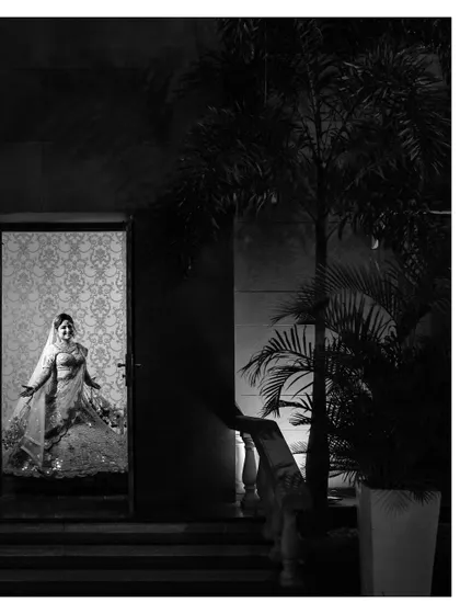 An artistic black and white portrait of the bride, seen through a doorway. The dramatic lighting and composition create a sense of anticipation and mystery.