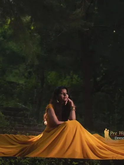 A moody, atmospheric shot of the mustard yellow trail gown in a forest setting. The fabric spreads out on the ground, creating a pool of warm color in the dim light.