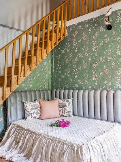 A whimsical bedroom nook designed under a wooden staircase. The custom-made daybed with a channel-tufted headboard is set against a lovely floral wallpaper, creating a magical little retreat.