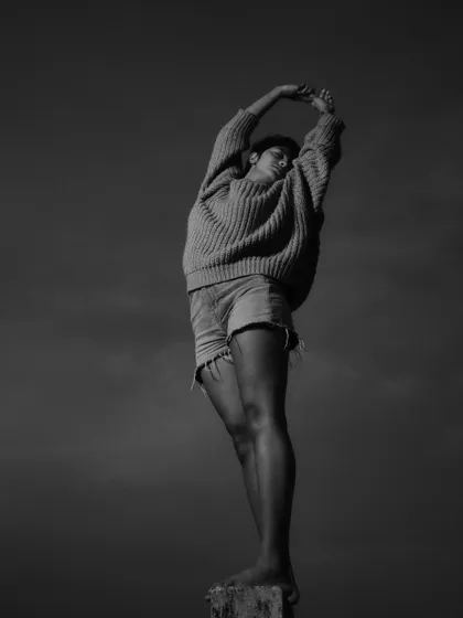A black and white conceptual portrait of a woman in a sweater and shorts, stretching towards the sky. The minimalist composition against a dark, open sky creates a sense of freedom and raw emotion.