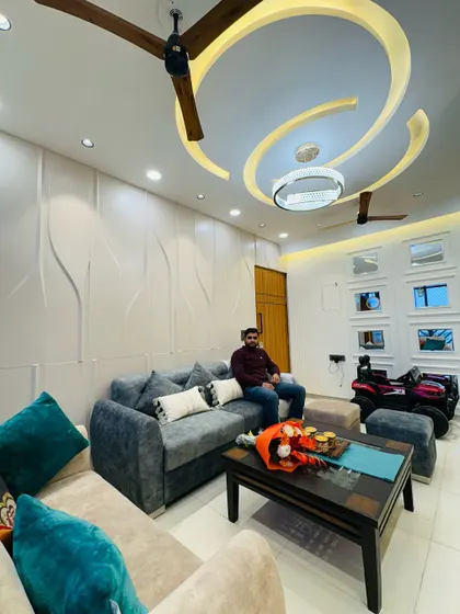 Here I am in a newly completed living room. The design features a 3D wave-patterned accent wall, a comfortable L-shaped sofa, and a false ceiling with a unique curved design, creating a modern and dynamic space.