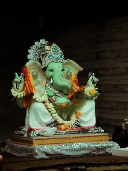 A beautifully crafted green Ganesh idol being worshipped in a pandal before the procession begins, with the warm light of an aarti flame nearby.