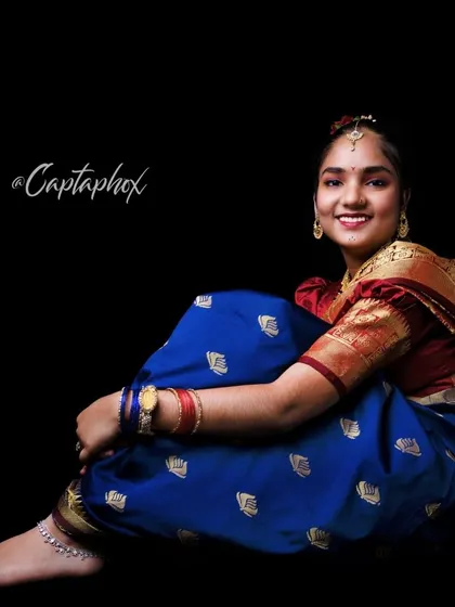 A full-body seated portrait that beautifully displays the drape and design of a traditional blue saree. The relaxed pose and warm smile create an inviting and elegant photograph.