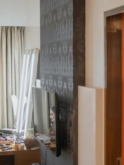 A creative getting-ready shot. The bride is seen applying makeup in the mirror, while the groom is captured getting ready in a separate room, visible through a doorway.