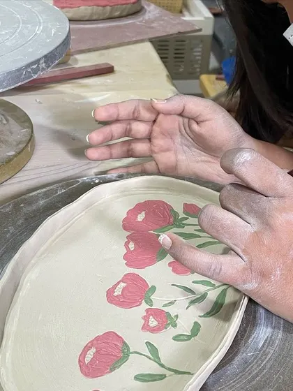 A student carefully paints delicate pink flowers onto their platter, a design that will become permanent and vibrant after glazing and firing.