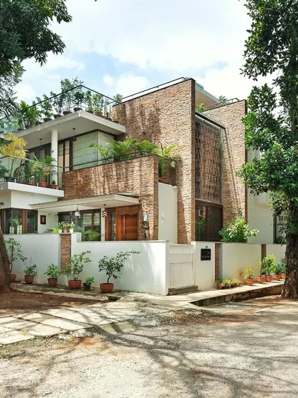 A street view of the completed Twin Courtyard House, nestled among mature trees. The design uses natural stone and prominent green balconies to blend in with its verdant surroundings.