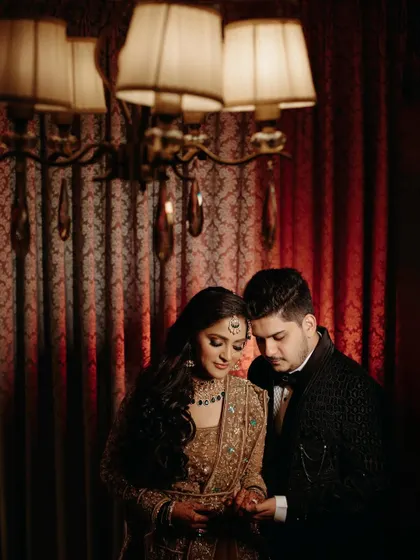 The couple examines the bride's ring in a quiet moment together. The dramatic, warm lighting and rich red backdrop create a moody and romantic atmosphere, perfect for capturing intimate details.