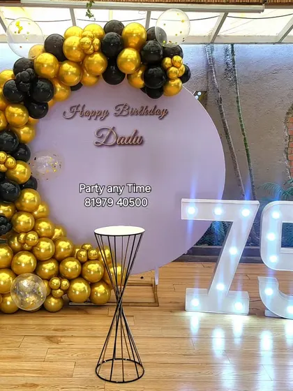 An elegant 75th birthday celebration with a black and gold balloon arch on a lavender backdrop, complete with large light-up marquee numbers.