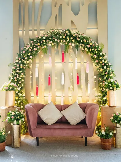 The main seating area for the Mehendi, featuring a plush pink sofa set against a modern backdrop with a floral arch and hanging tassels.