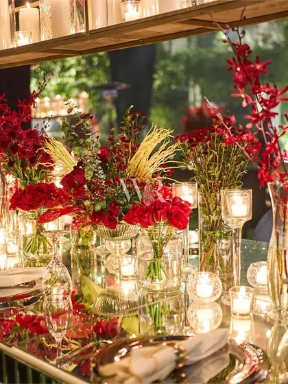A mirrored dining table is densely packed with red floral arrangements in clear glass vases, interspersed with candles. The overhead candle installation adds another layer of warm, flickering light.