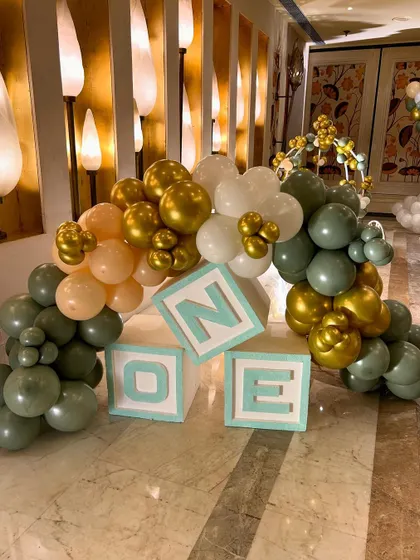 Light up "ONE" blocks surrounded by a garland of sage green, gold, and peach balloons, used as a decorative accent for the twins' first birthday party.