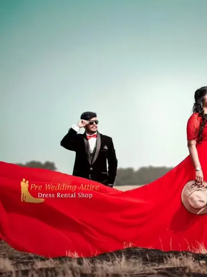 A stylish, editorial-style shot of a couple in a field. The woman's red trail gown billows in the wind, creating a high-fashion look.