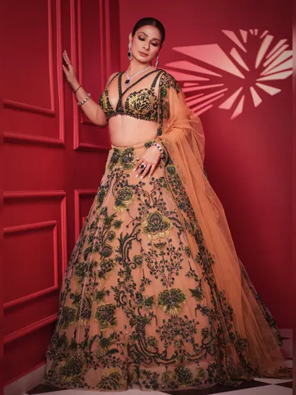 A dynamic pose against a bold red background. The contrast makes the green and peach embroidery of the lehenga pop, creating a visually striking image.