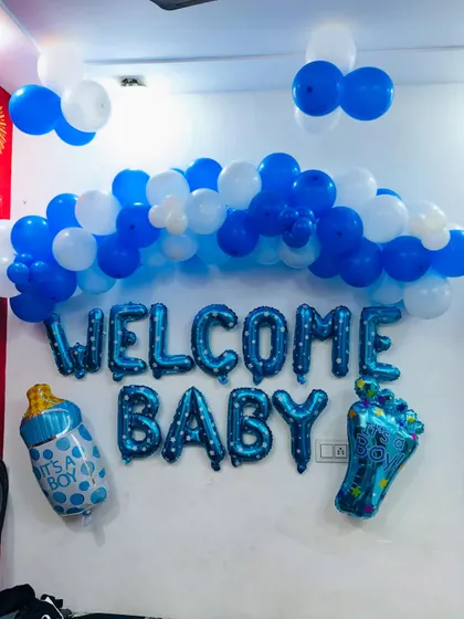 A 'Welcome Baby' wall decoration with a blue and white balloon arch and foil balloons including a baby bottle and a baby foot.
