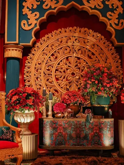 A close-up of the photo corner, highlighting the massive ornamental gold medallion and the rich textures of the backdrop. The space is filled with lush red floral arrangements to complete the regal look.