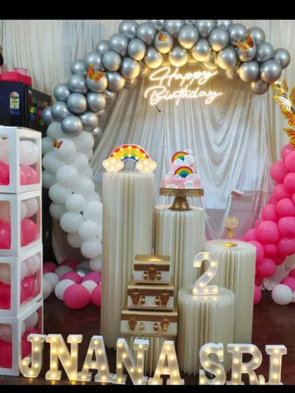 A cute second birthday party with a pink and silver balloon arch. The setup includes 'BABY' blocks and a rainbow cake topper.