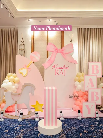 A dedicated photo booth with the baby's name, "Kaashvi Raj." The setup included a rocking horse, a rattle prop, and "BABY" blocks, all in a soft pink and white palette.
