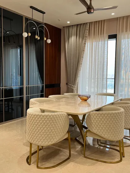 A luxury dining room featuring a large marble table and quilted cream-colored chairs with gold-finished frames. The smoked glass wardrobe panels in the background add depth and a touch of modern glamour.