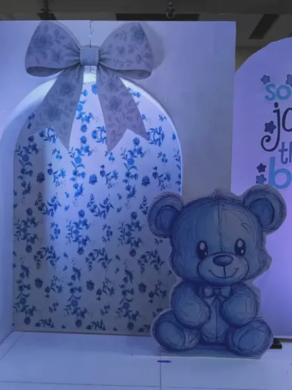A close-up of a baby boy shower backdrop. This setup features a large gift box design with a floral pattern, a cute teddy bear cutout, and the phrase "so much joy in this little boy".