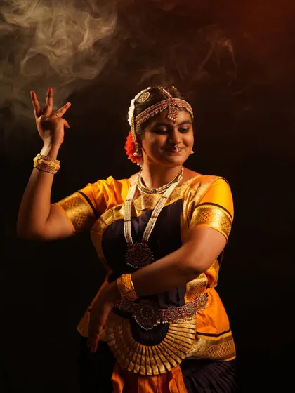 A close-up of a dancer in a traditional Bharatanatyam costume, highlighting the intricate temple jewelry, headpiece, and expressive hand gestures.