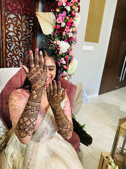 A playful bride peeking through her hands, which are covered in a beautiful floral and jaali design.