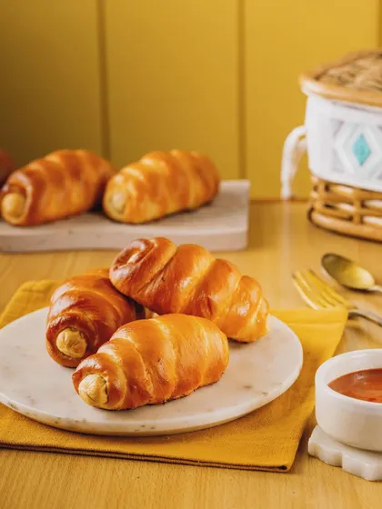 Golden-baked sausage rolls arranged on a marble plate against a warm yellow background. The soft lighting and simple styling highlight the buttery sheen of the pastry, making them look incredibly appetizing.