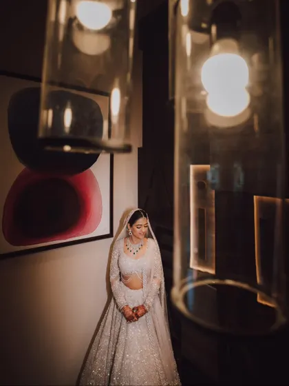 A bride standing under modern light fixtures, the warm light complementing her elegant white lehenga.
