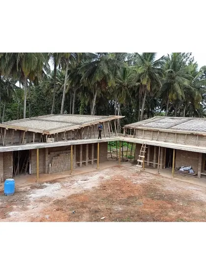 An aerial view during the construction phase, showing the two building blocks arranged around the central courtyard. The structure is set against a backdrop of coconut groves, highlighting its integration into the local landscape.