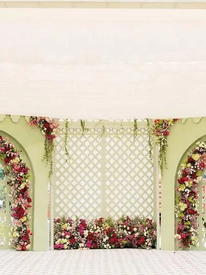A charming and colorful photo backdrop for a Parisian-themed Haldi, with two floral arches set against a pastel green wall with lattice details.
