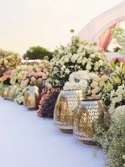 The wedding aisle is lined with lush arrangements of pastel flowers and ornate golden lanterns, creating a beautiful and luxurious path for the bride.