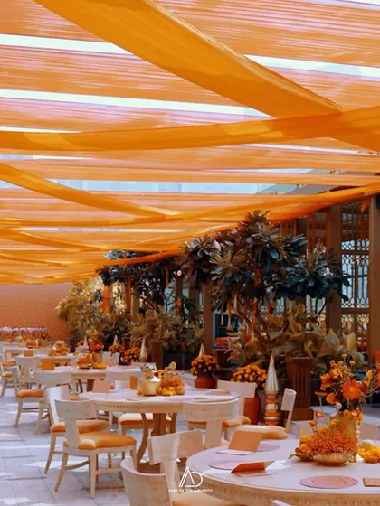 An outdoor dining setup for a Mamera ceremony, with tables adorned with orange floral centerpieces under a canopy of flowing orange drapes.