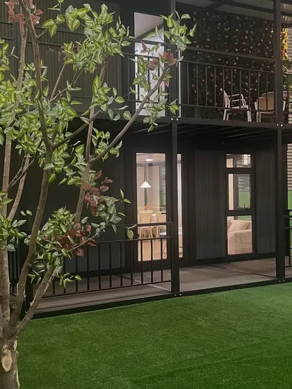 A side view of the two-story container home, highlighting the wrap-around porch and balcony. The black steel frame provides a strong visual contrast to the surrounding greenery.