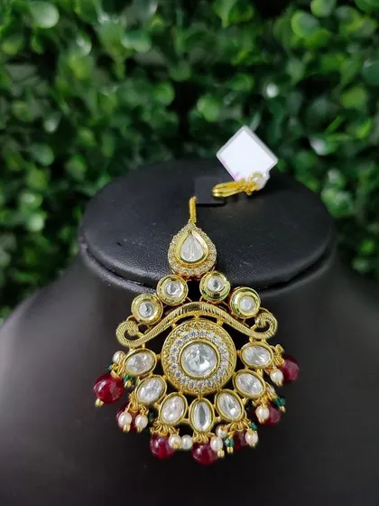 A beautiful kundan maang tikka with a central clear stone, surrounded by smaller stones and red bead drops.