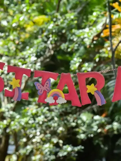 A personalized "Sitara" name banner with rainbows and butterflies, hanging outdoors for a jungle or fairy-themed birthday party.