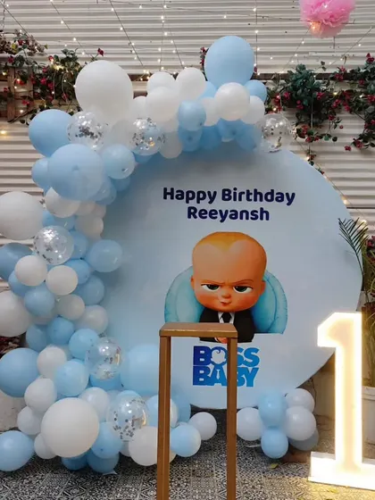A fun Boss Baby first birthday party. This outdoor setup includes a custom backdrop, a blue and white balloon arch, and a light-up number '1'.