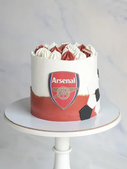 For an Arsenal football club fan, this cake features the team's logo and colors. The design includes a soccer ball pattern on the side and is topped with red and white frosting swirls.