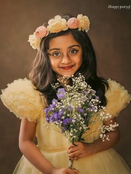 A girl in a yellow gown and floral crown holds a bouquet of purple and white flowers. Her sweet smile makes this a heartwarming portrait.