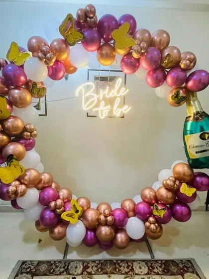 This glamorous balloon ring combines rose gold, magenta, and white balloons with gold butterfly accents. A 'Bride to be' neon sign and a large champagne bottle balloon make it clear that it's time to celebrate.