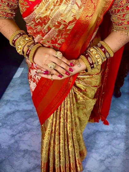 A close-up of a South Indian bride's hands, adorned with henna and bangles, against the backdrop of her perfectly draped red and gold Kanchivaram.